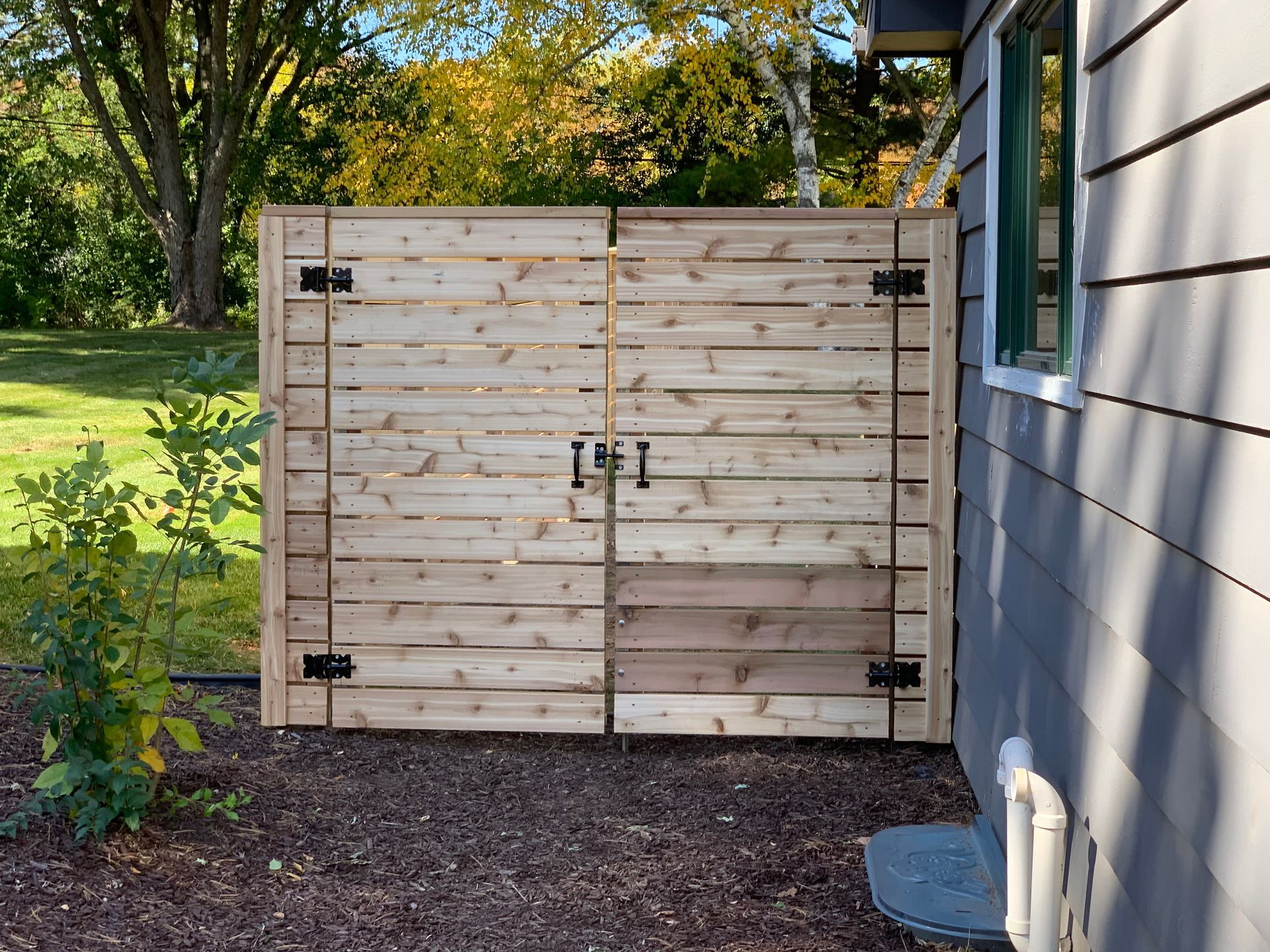 Wooden privacy fence with double doors against a gray house, on grass near a tree.