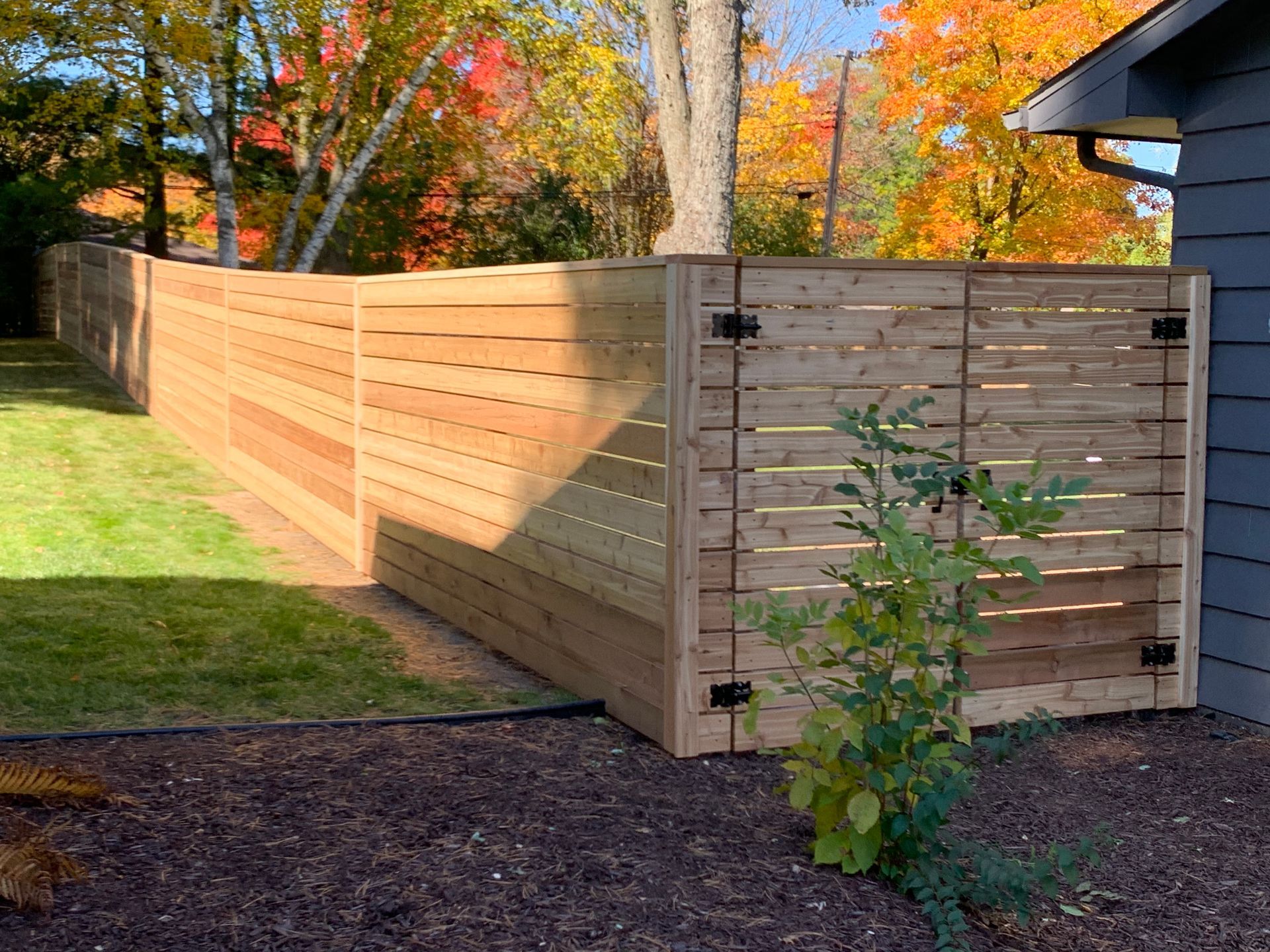 Wooden horizontal slat fence in a yard with fall foliage in the background.