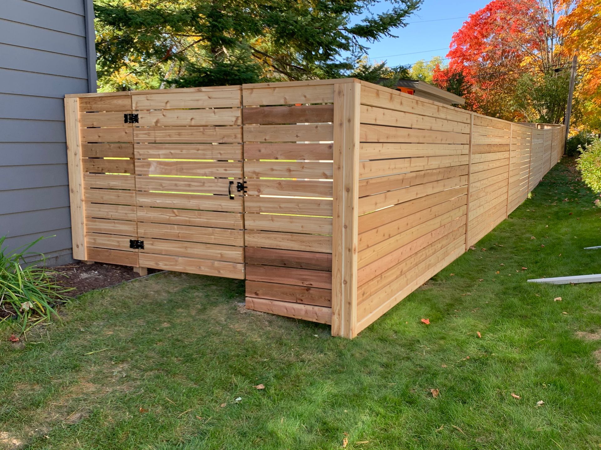 Wooden fence with gate, bordering a green lawn, next to a gray house. Fall foliage in background.