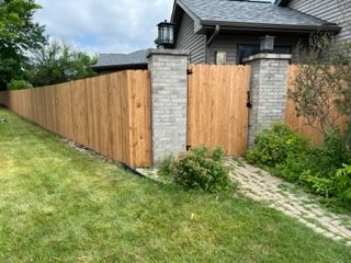 Wooden fence with brick columns and gate, beside a house with a green lawn and paved path.