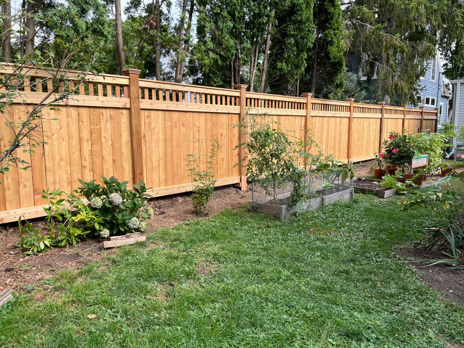 Wooden fence bordering a backyard garden with green grass, flowers, and trees.