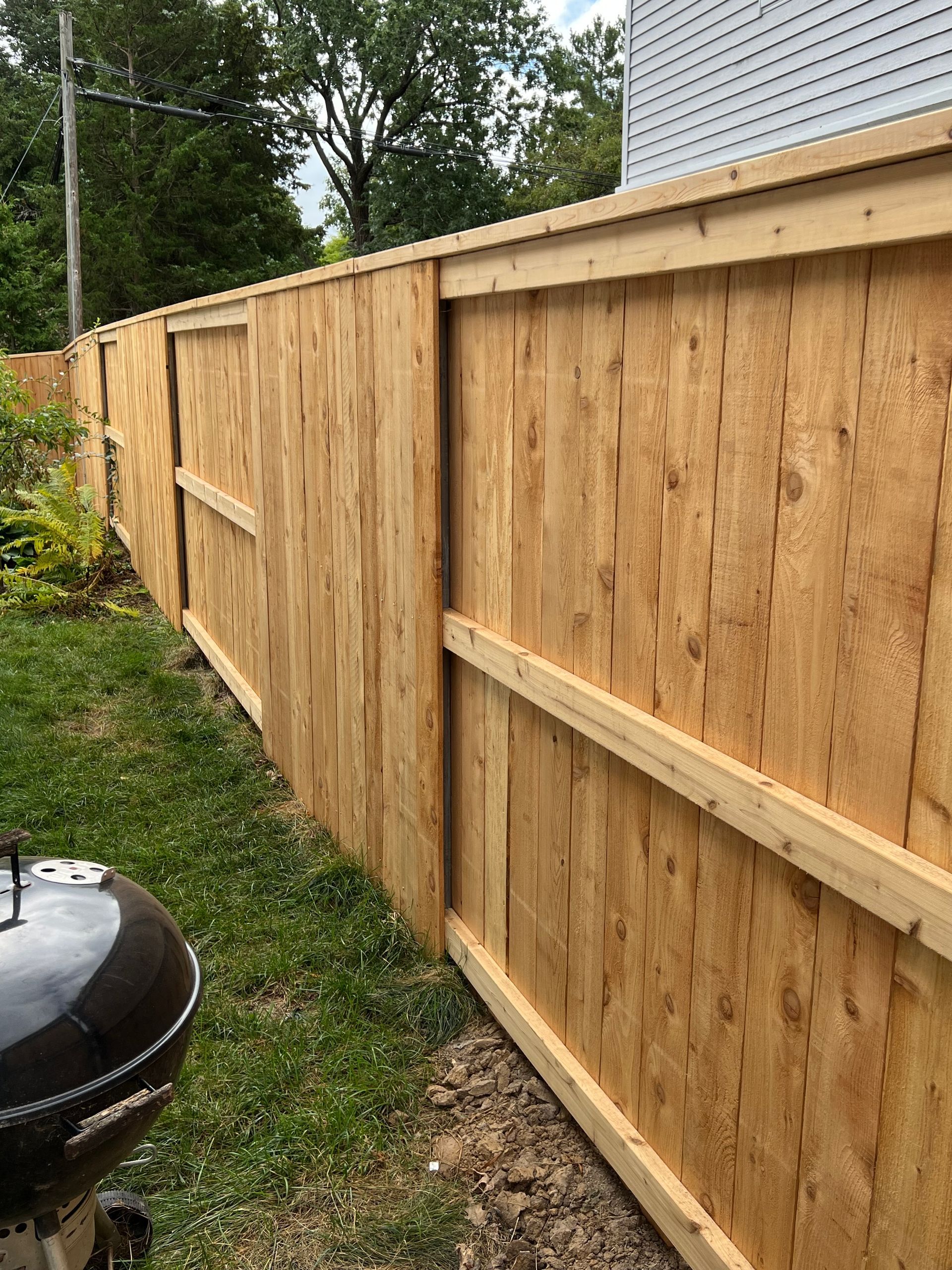 Wooden fence on green grass, with a dark grill in the foreground, and a house and trees in the background.