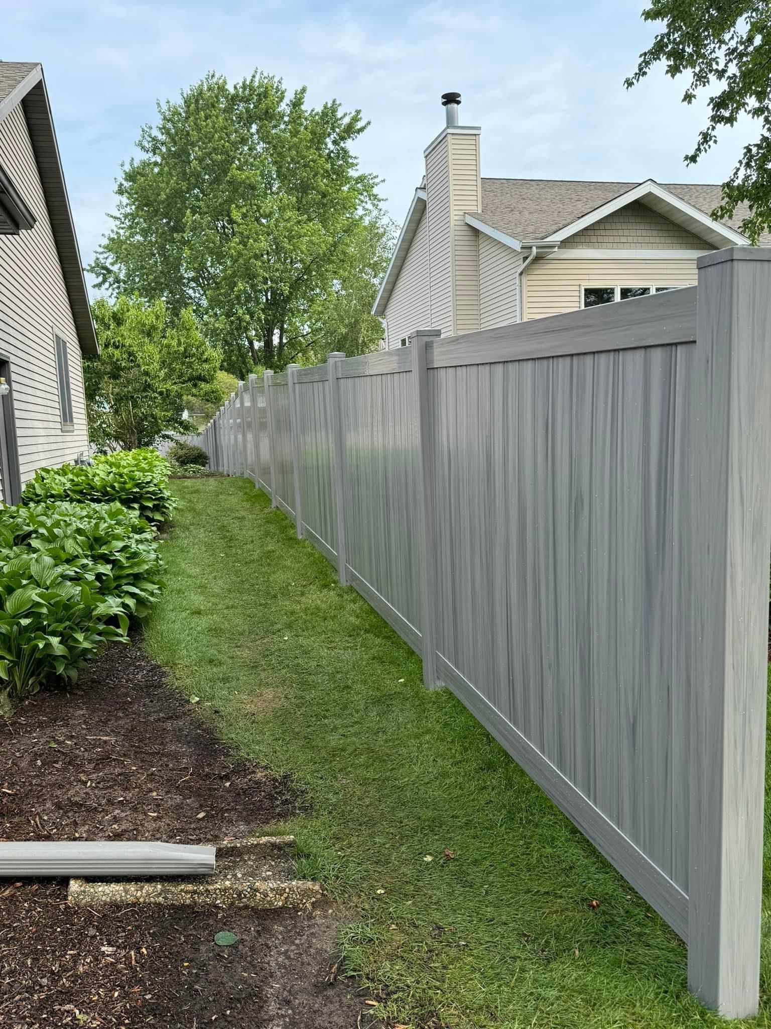 Grey wooden fence along a grassy yard, beside a house and landscaping.