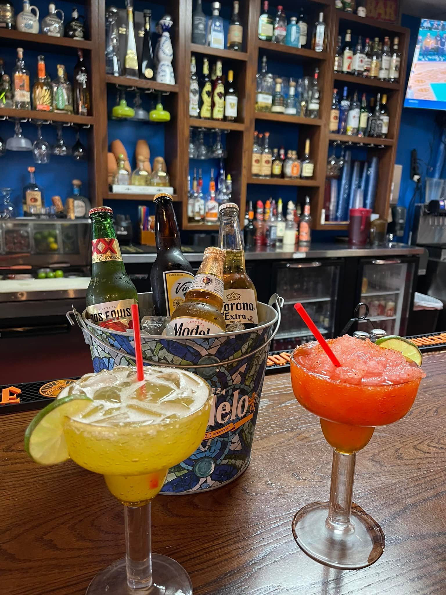 Two margaritas are sitting on a wooden table in a bar.