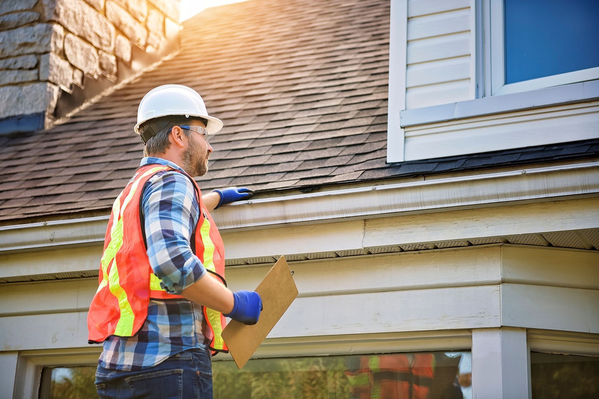 Worker in safety vest and hard hat inspecting a house roof with a clipboard