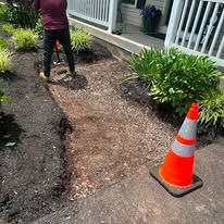 Driveway sidewalk repair with exposed soil, a worker, and an orange traffic cone beside a porch.