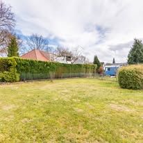 Grassy backyard with hedges, a house, and a blue shed under a cloudy sky