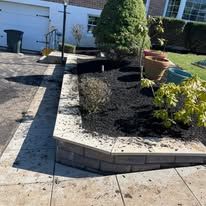 Raised garden bed with dark mulch, shrubs, and a tree beside a paved patio near a house