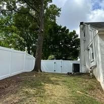 Backyard with white fence, large tree, grassy yard, and gray house under a cloudy sky