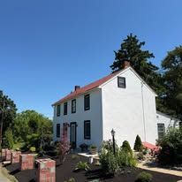 White two-story house with red roof and black-trimmed windows, surrounded by greenery under a clear blue sky