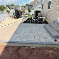 Backyard patio with gray pavers, outdoor chairs, and a partially finished soil edge.