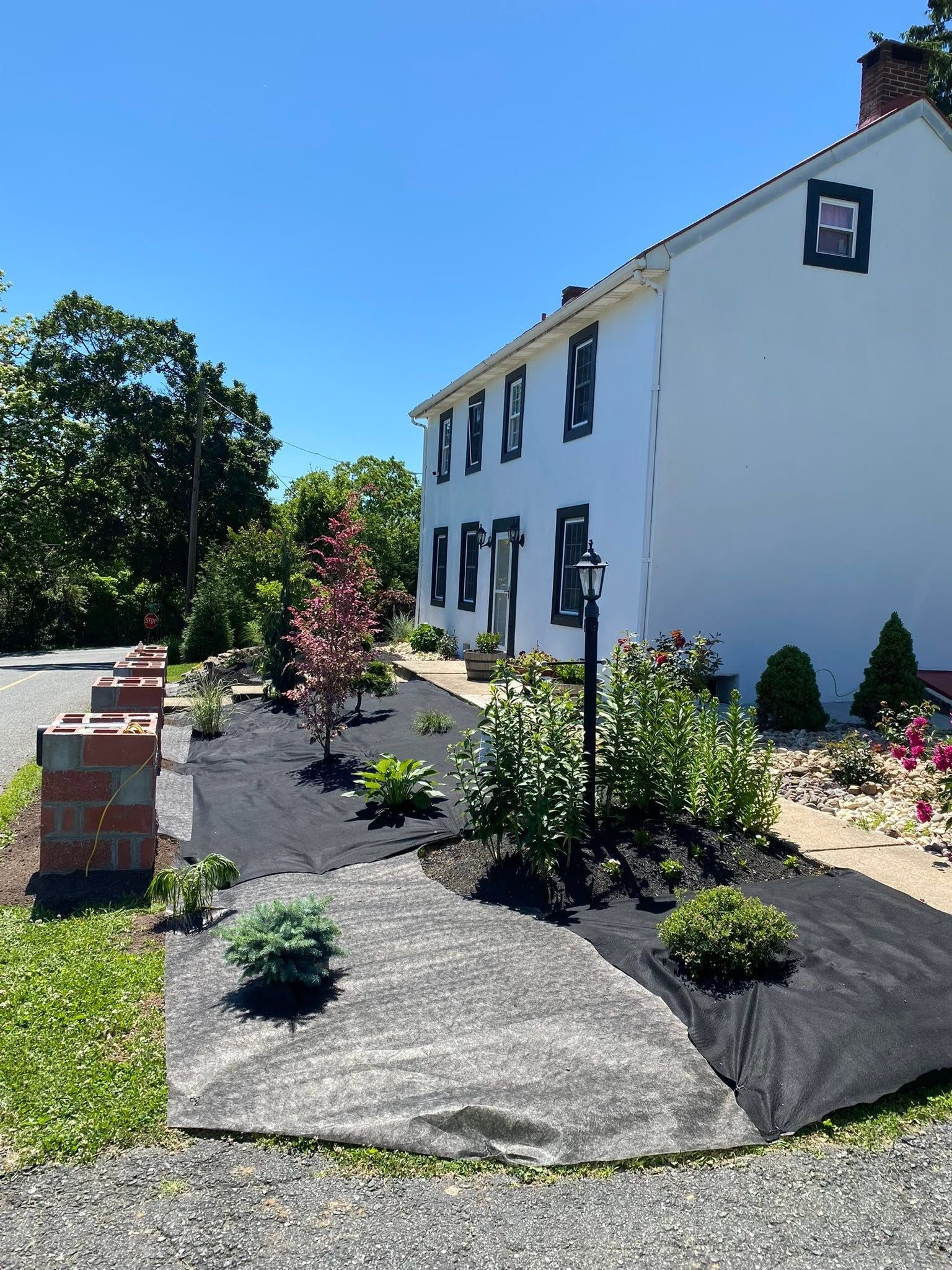 Front yard landscaping beside a white house with stone paths, shrubs, and brick planters under a blue sky
