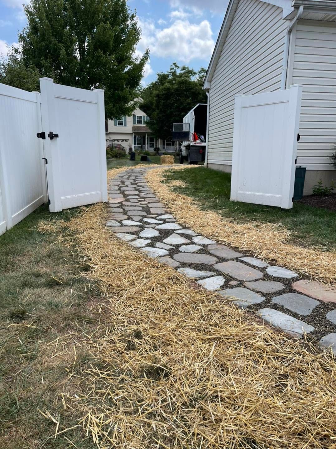 Stone path through open white gates beside a house, with dry grass and a tree under a partly cloudy sky