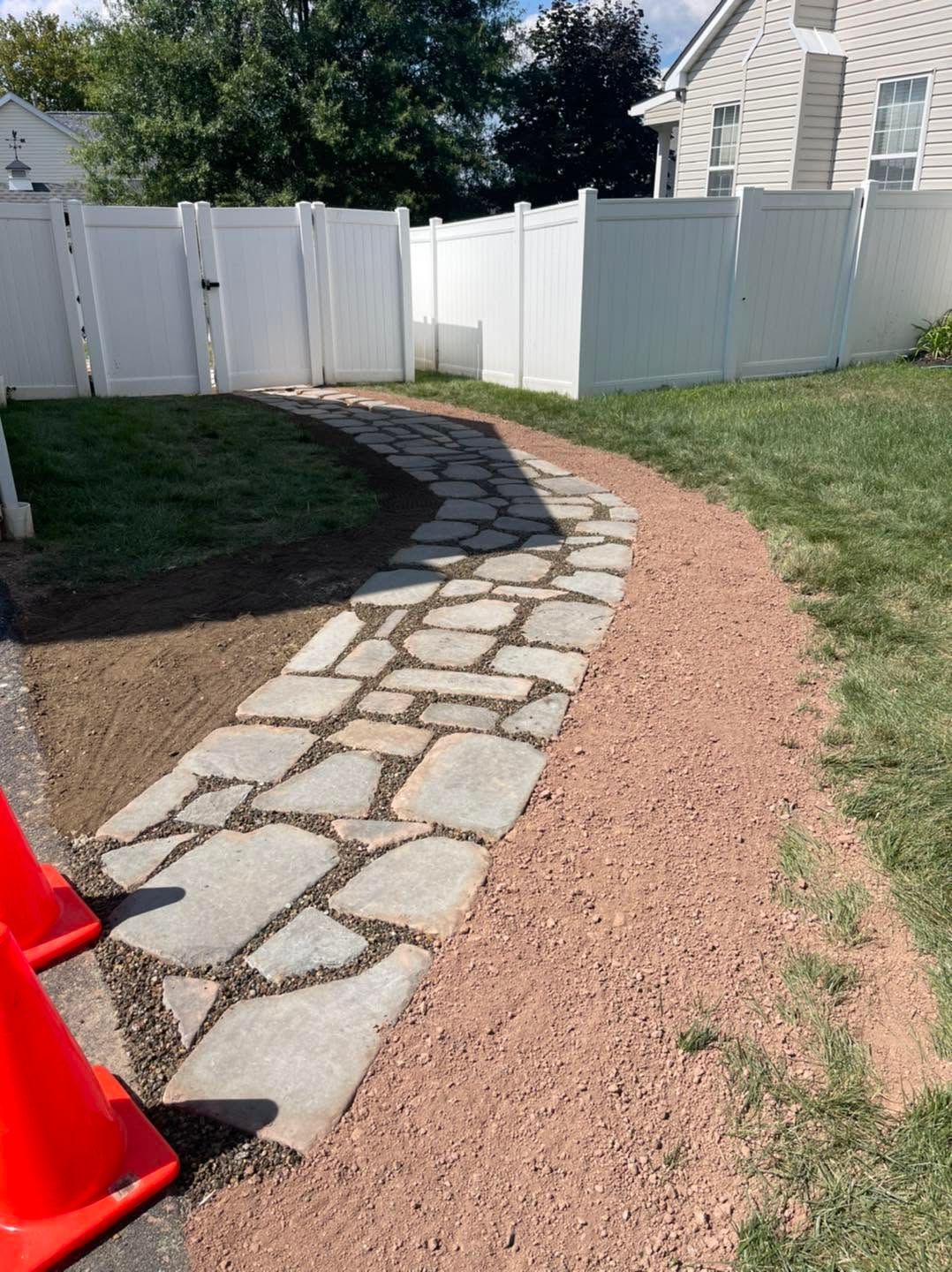 Curved stone walkway beside a white fence and house, with gravel edging and an orange cone.