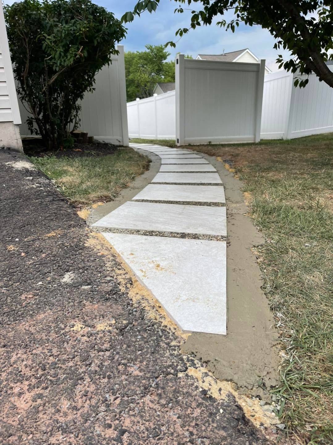 Curved concrete walkway leading to a white gate beside a grassy yard and trees