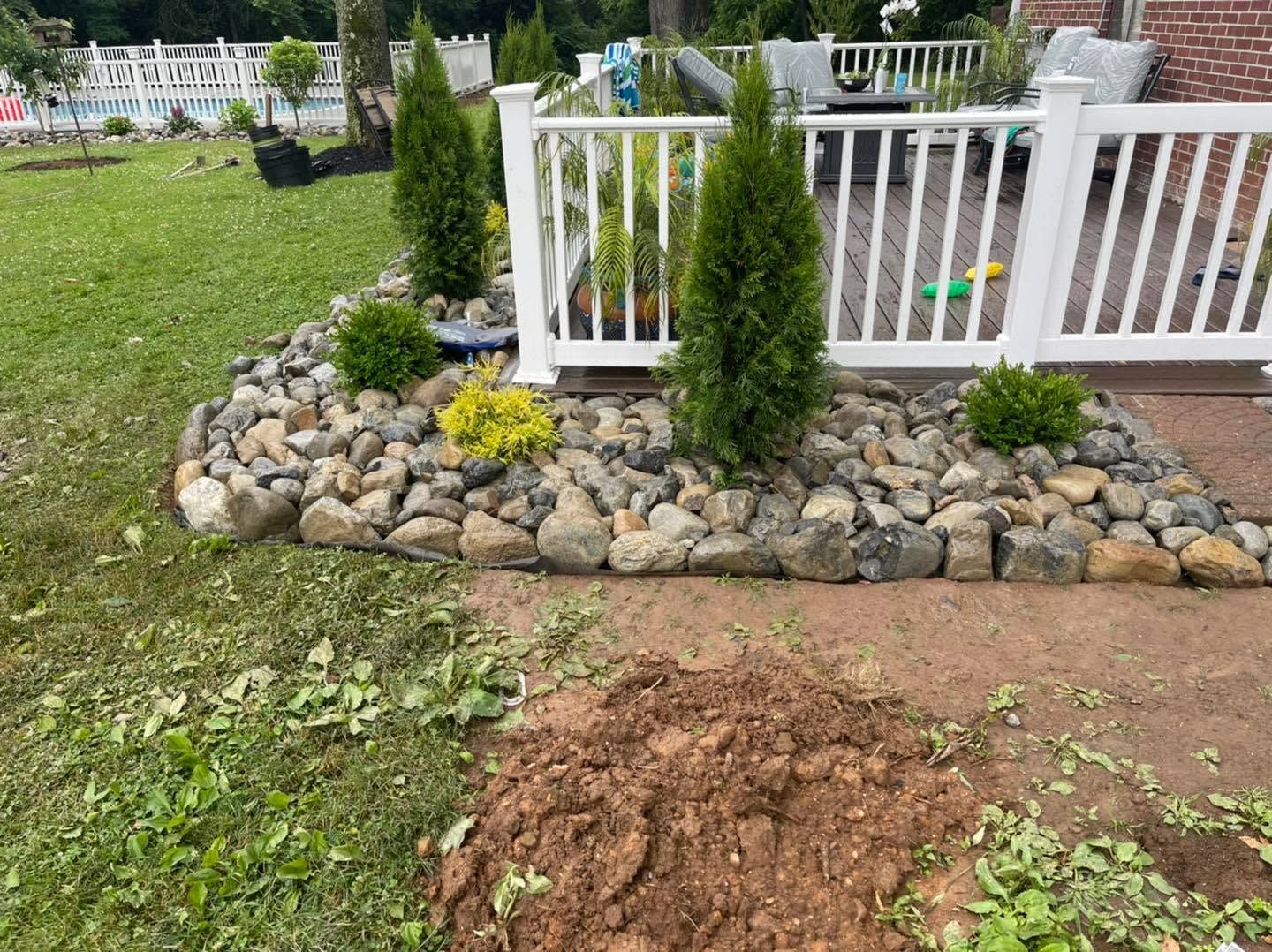 Rock-bordered garden bed with small evergreen shrubs beside a white deck on a grassy lawn.