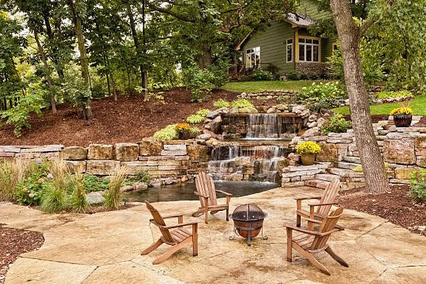 Outdoor patio with wooden chairs around a fire pit, overlooking a stone waterfall and wooded garden