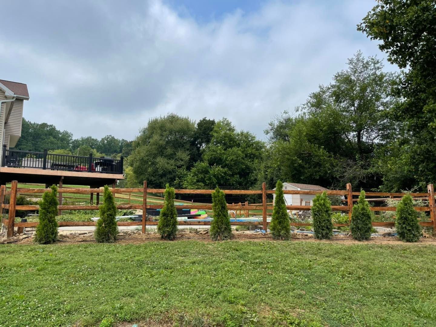 Grassy field with small shrubs, a wooden fence, and cloudy sky beside a house and trees