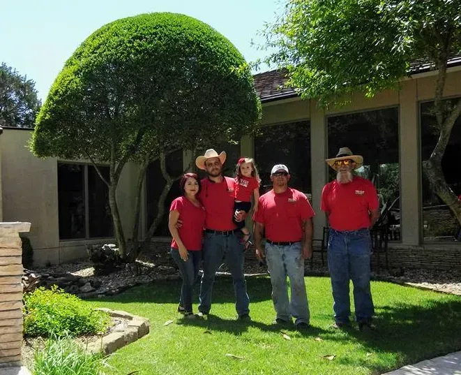 Five people in red shirts and cowboy hats pose outdoors in front of a tree and building.