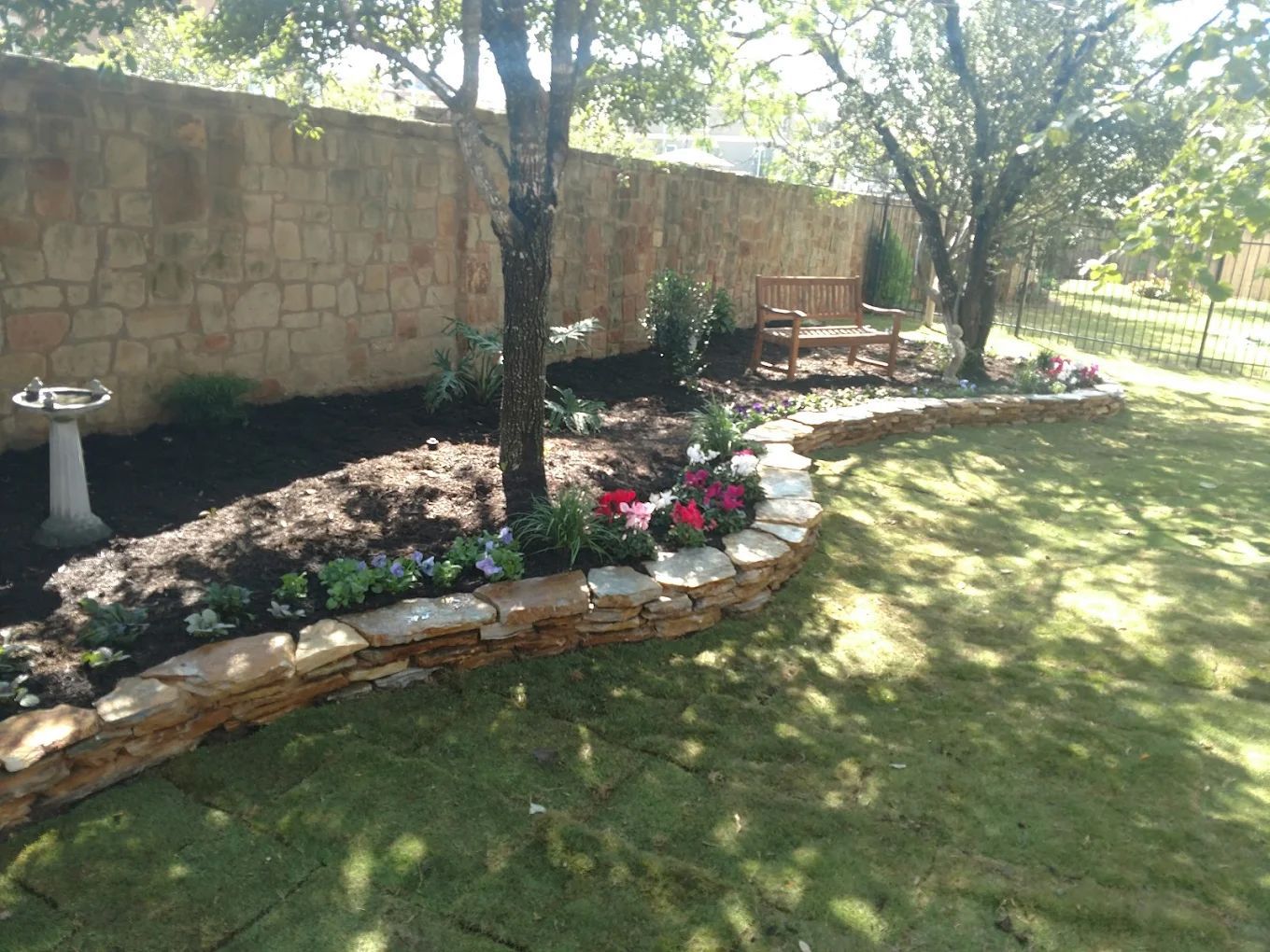 Stone-edged flower bed in a garden with a wall and birdbath, featuring flowers, trees, and a wooden bench.