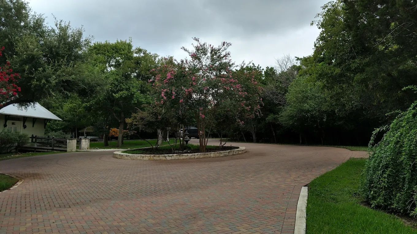 Brick driveway leading to a landscaped area with trees and a small building under an overcast sky.