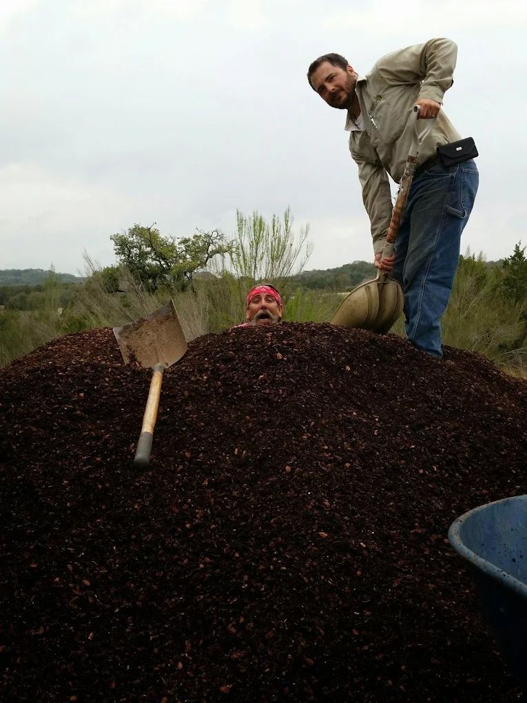 Man shovels dirt onto a pile, burying another person up to their head outdoors.