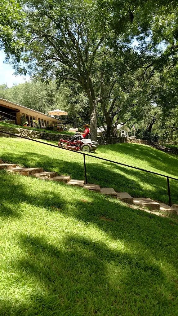 Lawnmower on a sloped green yard with steps, a house in the background, and large shade trees.