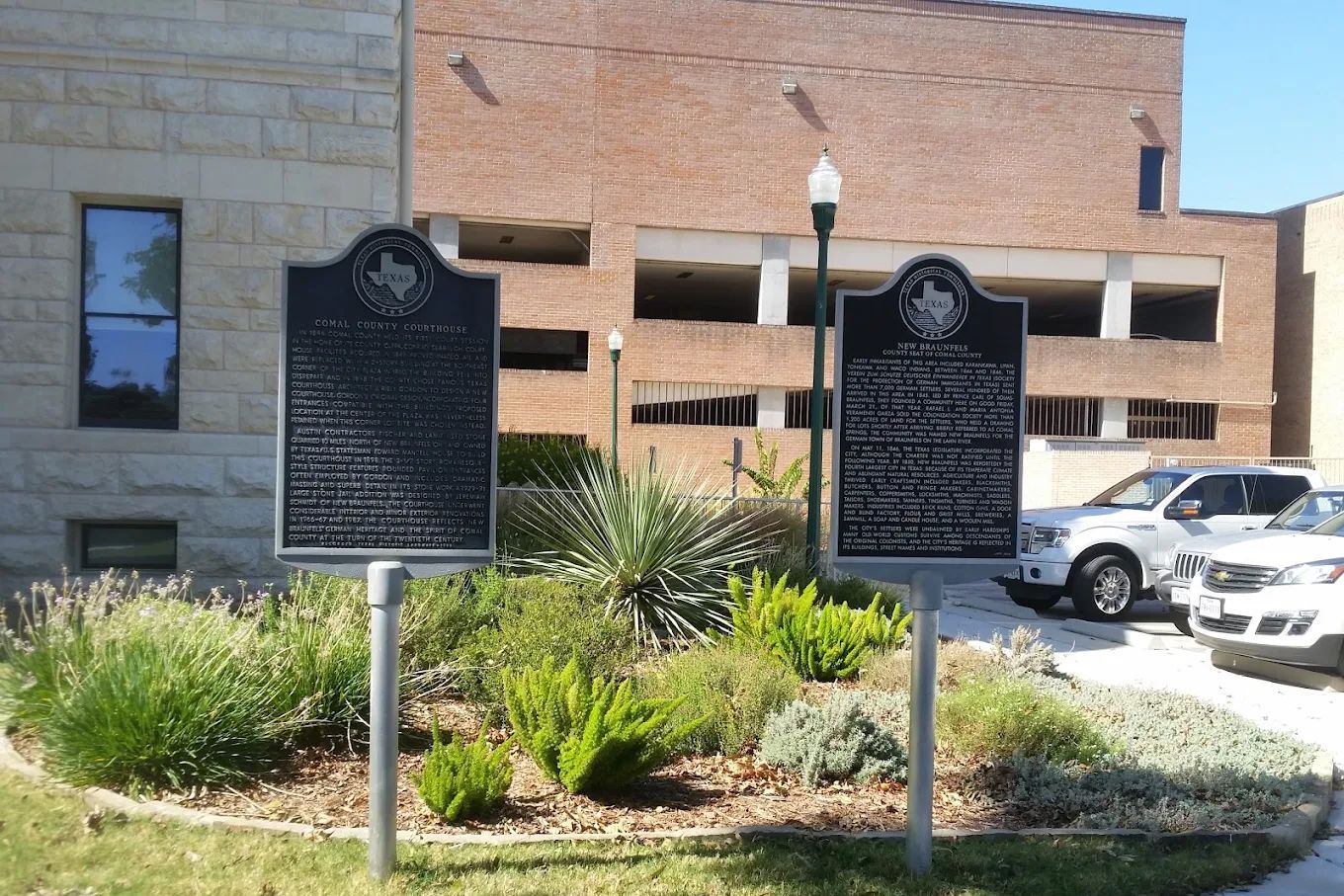 Two historical markers stand in a garden; brick building in the background; parked cars.