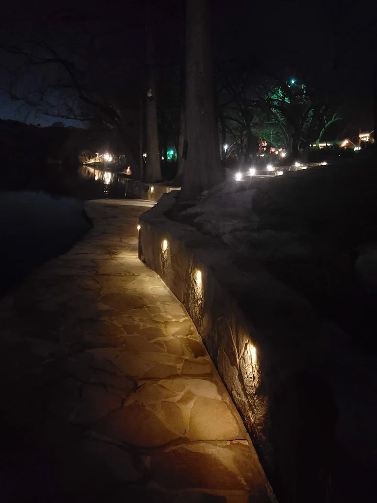 Stone pathway lit by small lights at night, curving toward a dark, illuminated landscape.