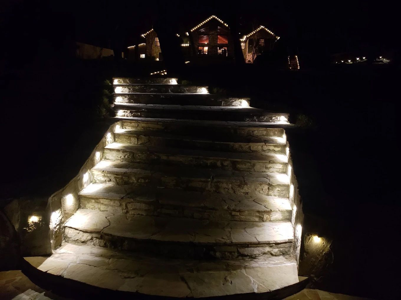Stone steps illuminated by lights lead to a house with Christmas lights at night.