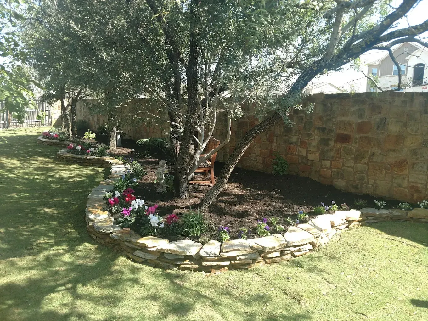 A stone-edged flower bed with colorful blooms encircles a tree, next to a stone wall, on a grassy lawn.