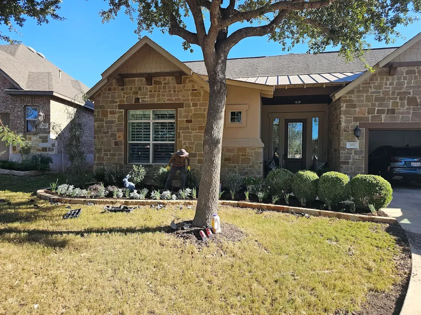 A stone-faced house with a tree in the yard. Green bushes line the flower beds. Blue sky overhead.