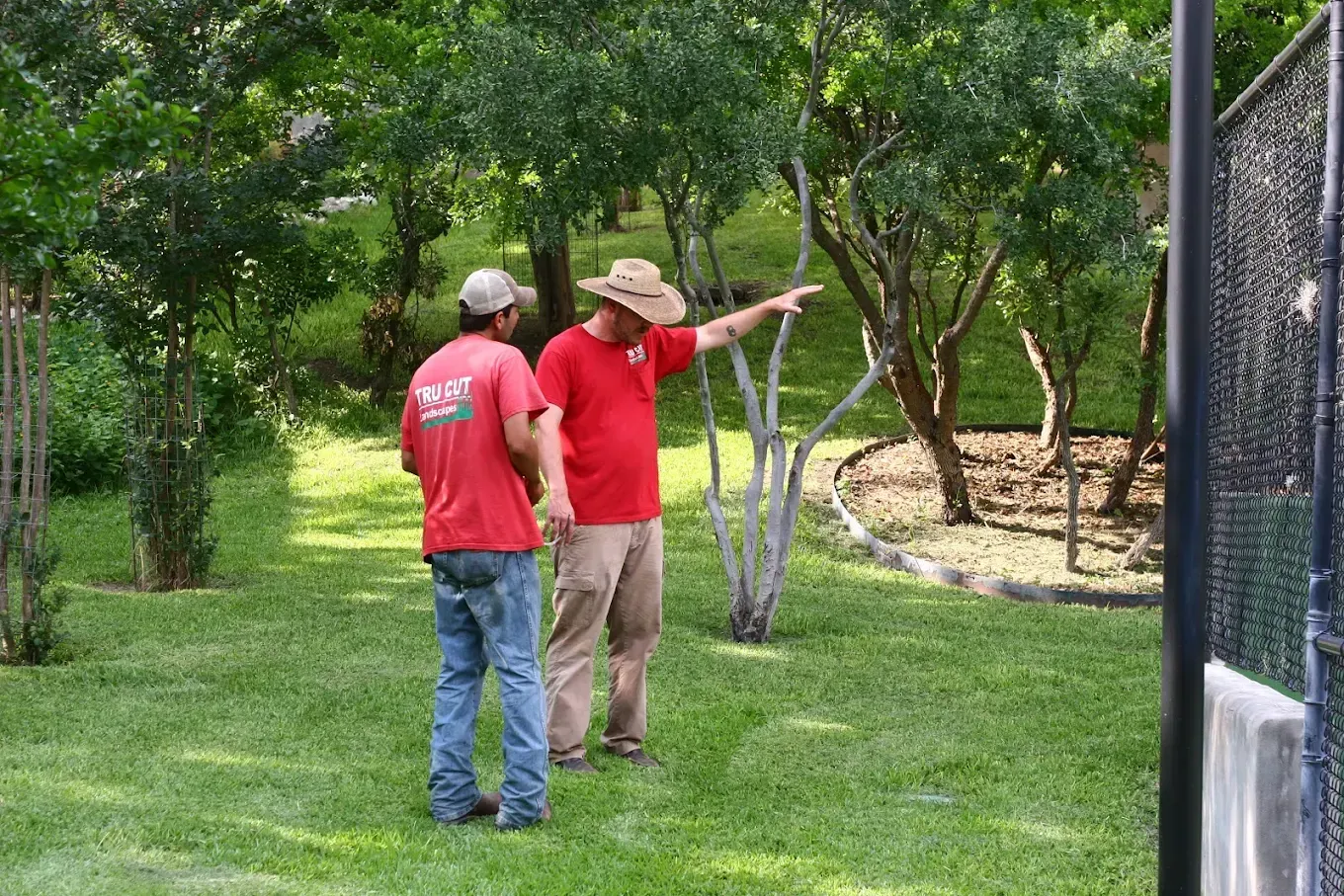 Two men in red shirts and khaki pants surveying a grassy area in a park, one gesturing.