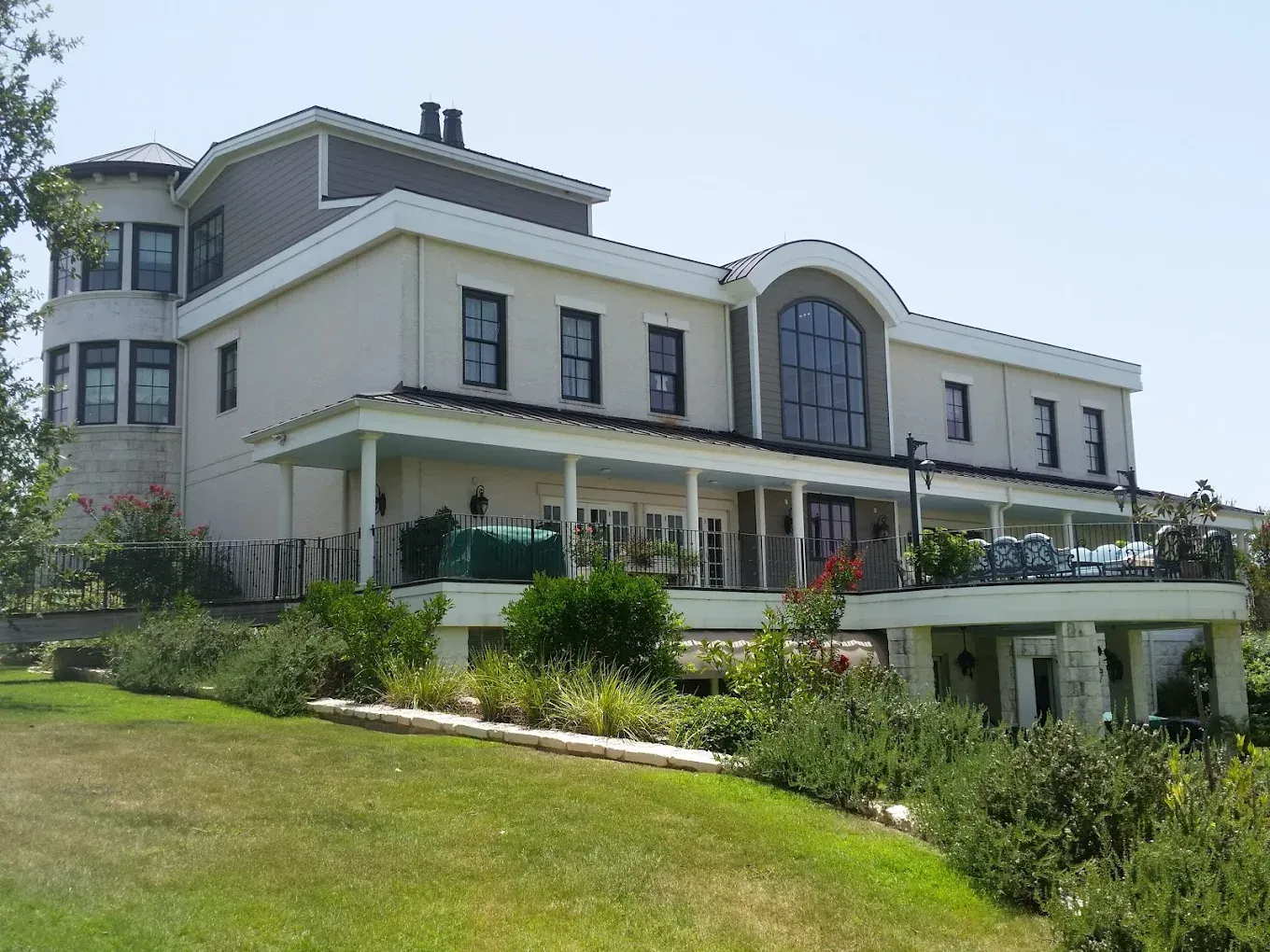 Large two-story beige house with a wraparound porch and landscaped yard under a clear blue sky.