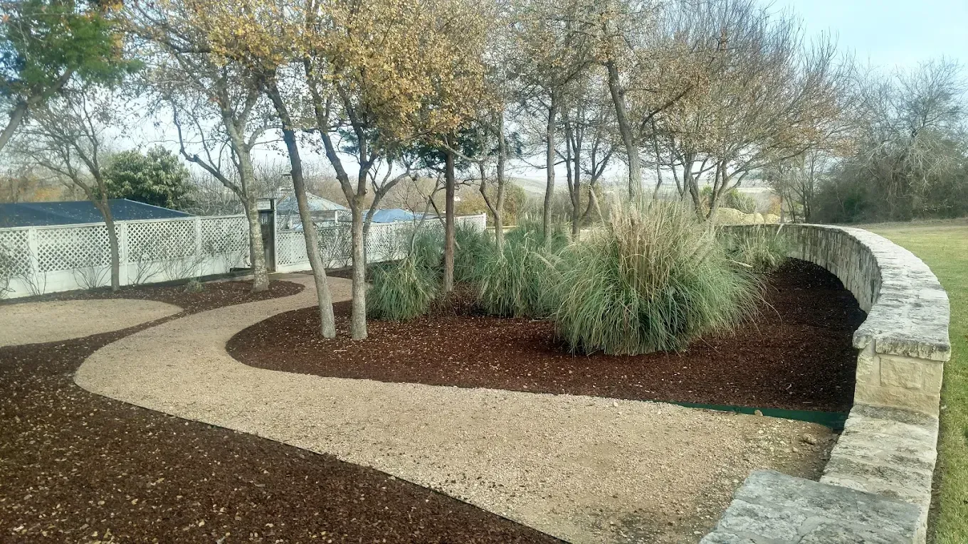 Winding gravel path bordered by brown mulch and a stone wall, leading through trees with bare branches.