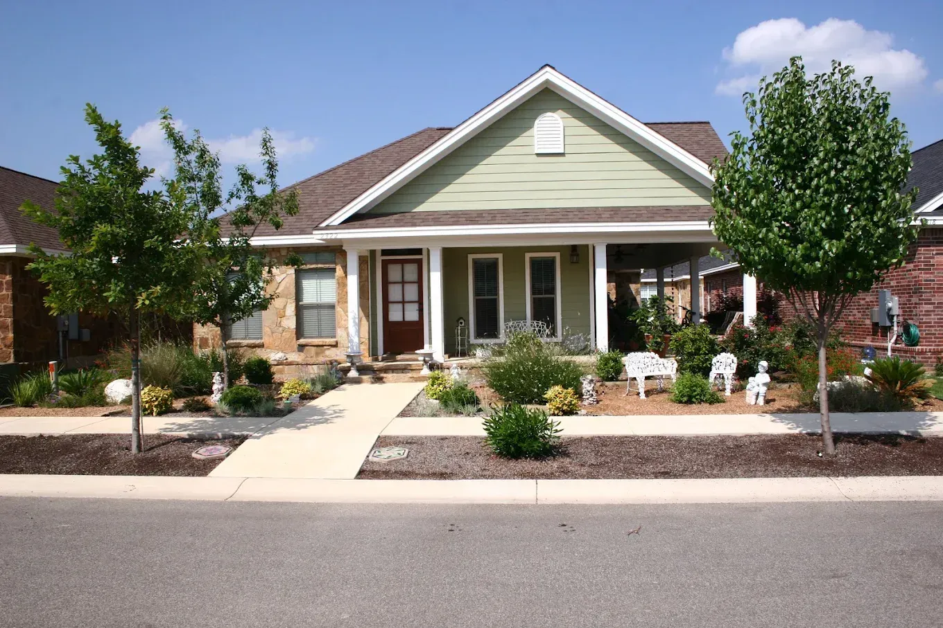 Single-story house with green siding, brown door, and porch. Landscaping includes trees, bushes, and mulch.