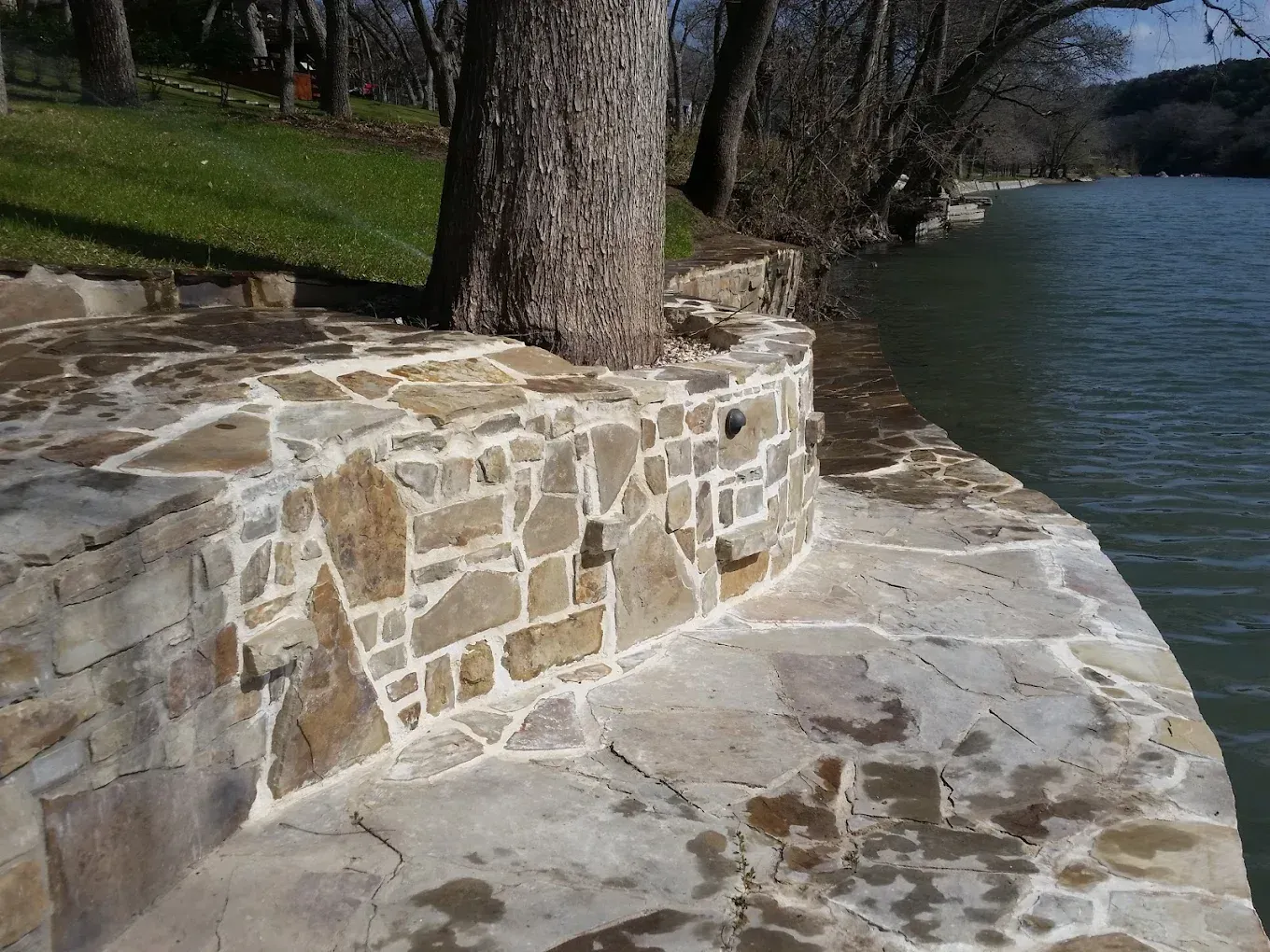 Stone patio and retaining wall by water, tree in center.