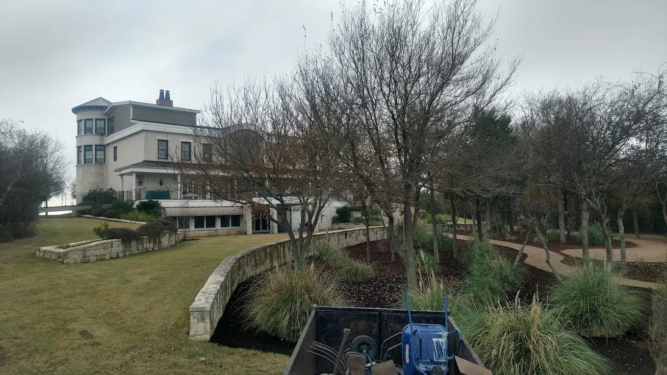 Large light-colored house with a turret, on a grassy hill. Trees and landscaping in foreground, overcast sky.