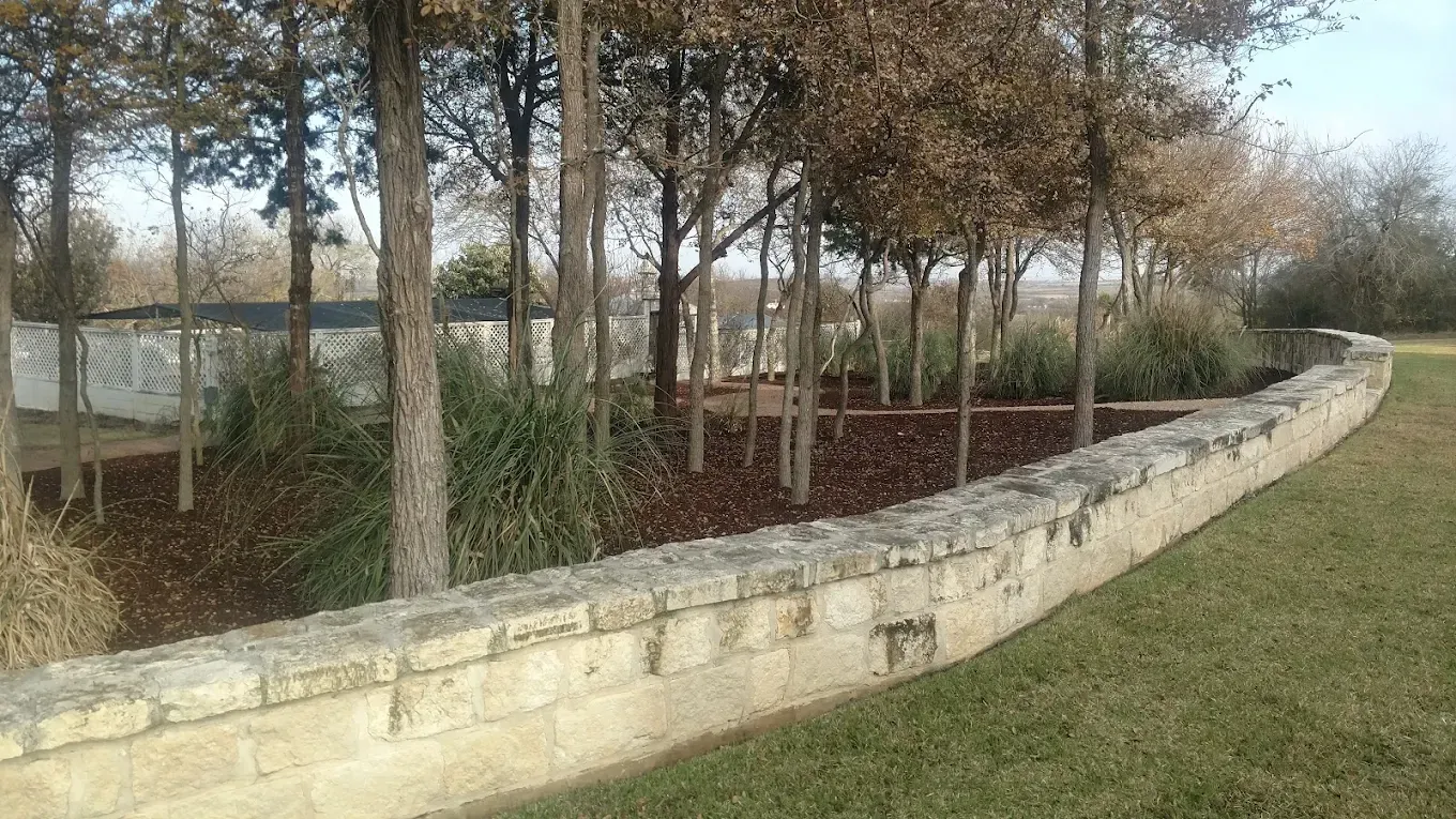 Low stone wall bordering a bed of trees and mulch in a grassy area.