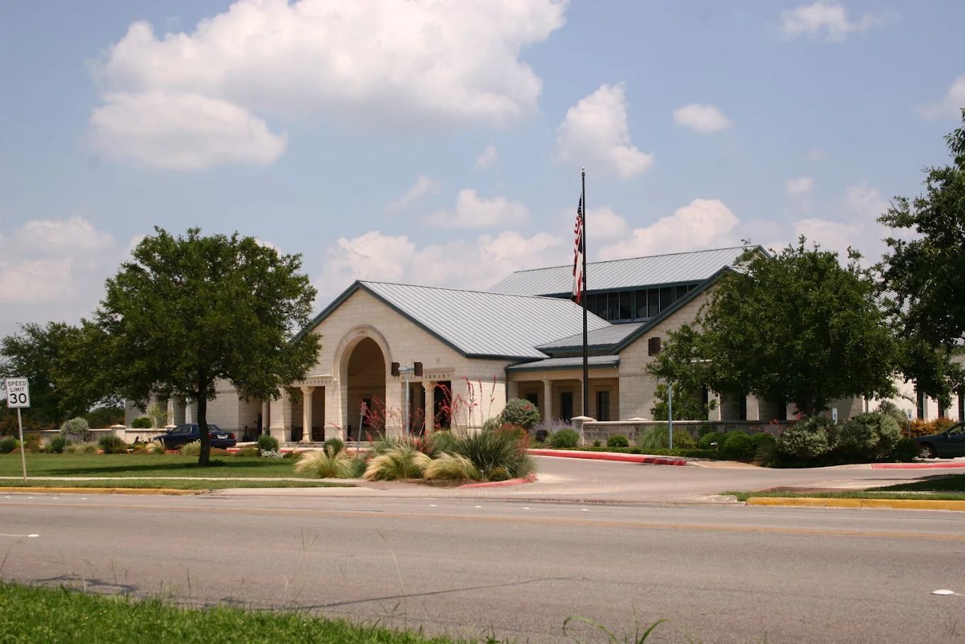Light-colored building with arched entrance, American flag, trees, and cloudy sky.
