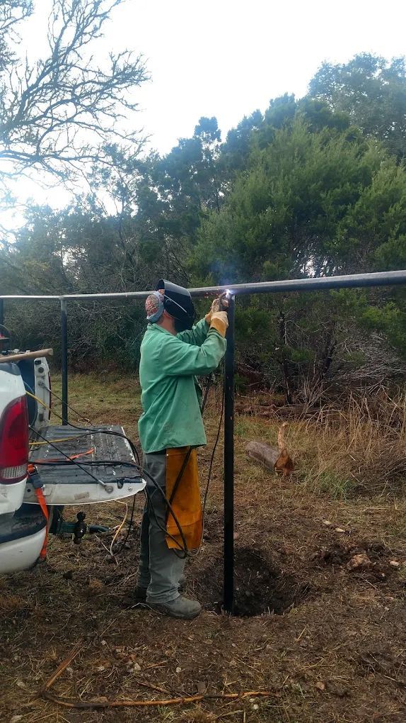 Person welds black metal fence in an outdoor setting, wearing safety gear. A truck is nearby.