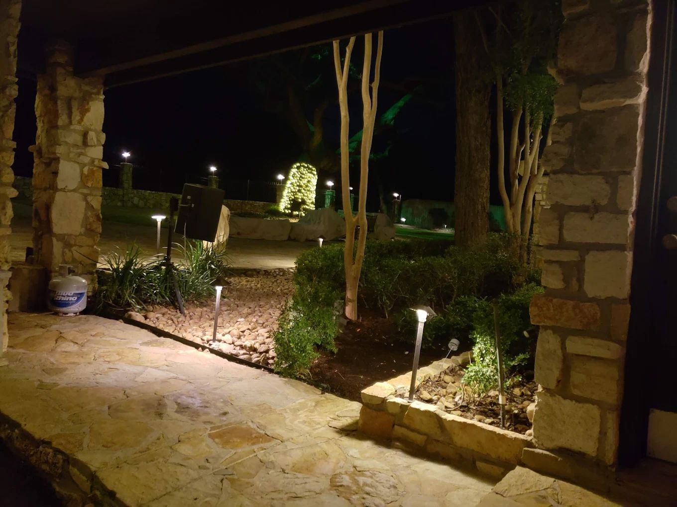 Night view of lit stone path, leading to a home's entrance, with landscape lighting illuminating the surrounding greenery.