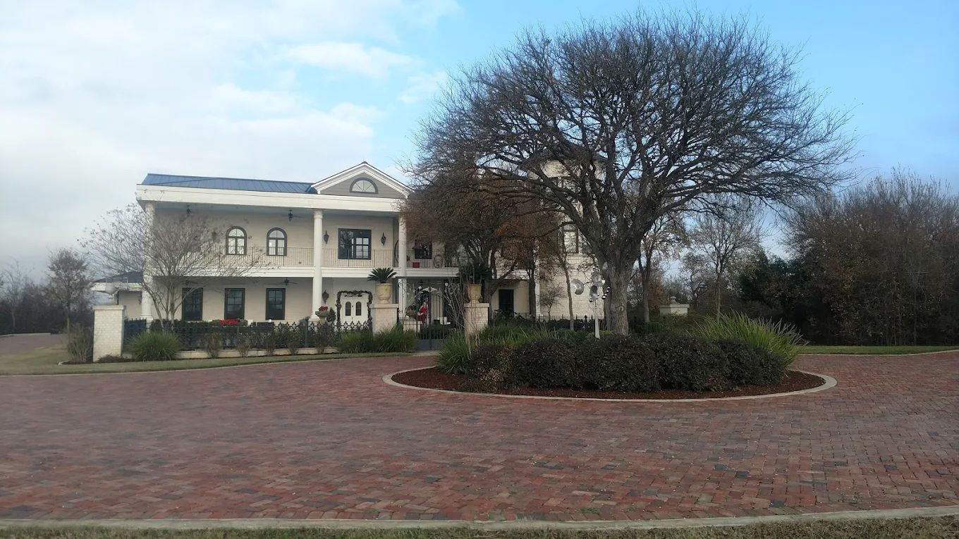 Two-story white house with brick driveway, a large tree, and a shrubbery border.