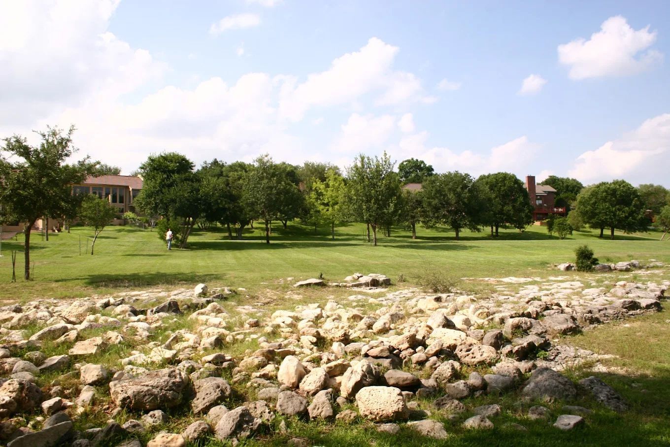 Green grassy field with scattered rocks in the foreground, trees, and buildings under a sunny sky.