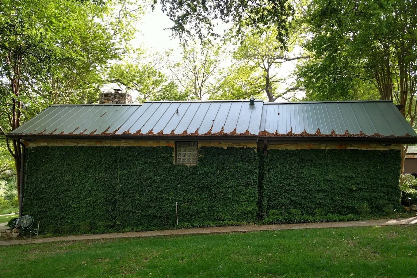 Green-covered building with a rusted metal roof, small window, and stone chimney, set against a backdrop of trees.