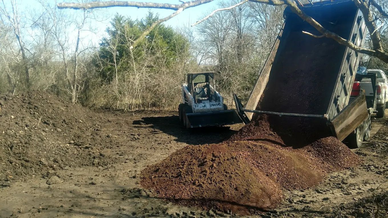 Skid steer unloading dirt from a trailer onto a muddy area near trees.