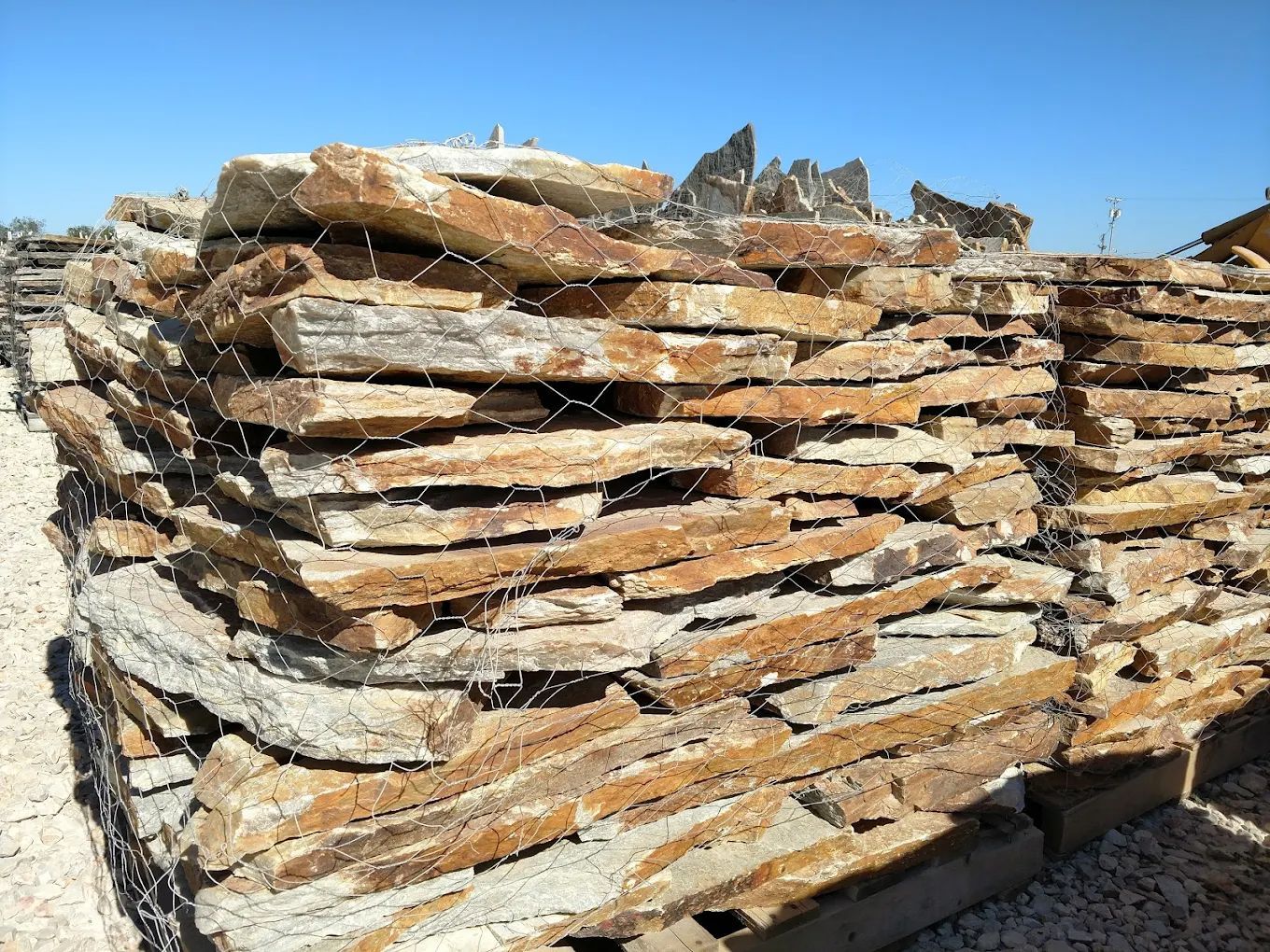 Stack of irregularly shaped brown and tan natural stone slabs bound with wire mesh, outdoors under a blue sky.