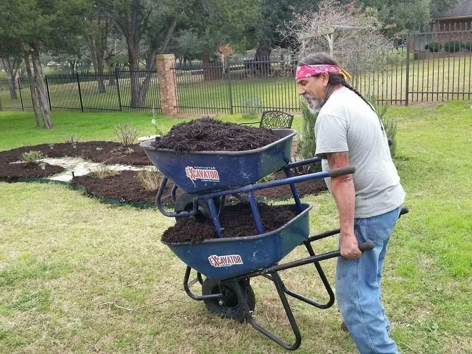 Man struggling to push two stacked wheelbarrows full of dark soil on a grassy lawn.