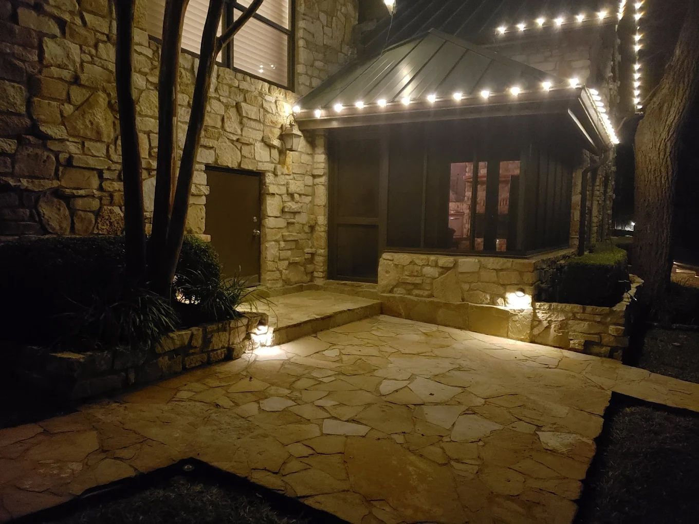 Stone building entrance at night, lit by string lights and spotlights. Brown door and stone patio.