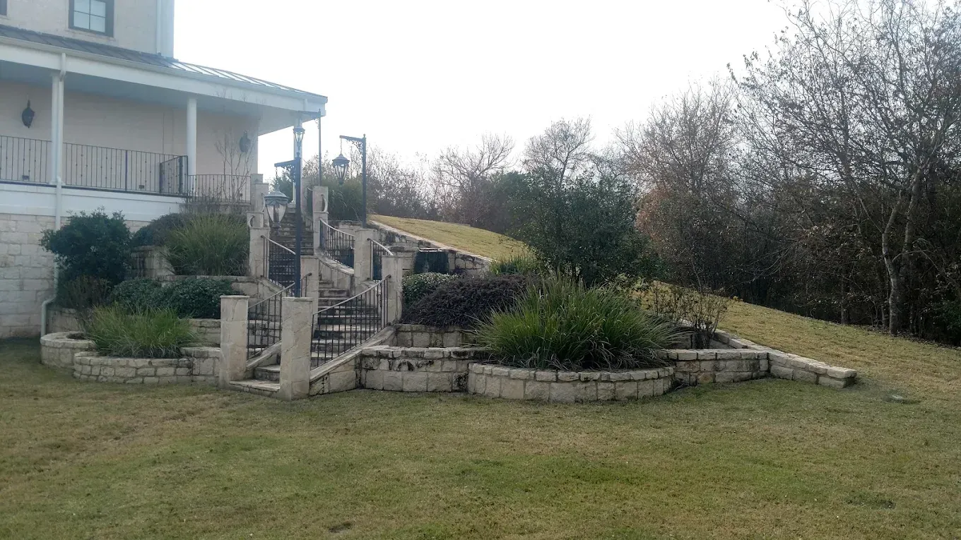 Stone steps and tiered planters leading up a grassy hill next to a house. Overcast sky.